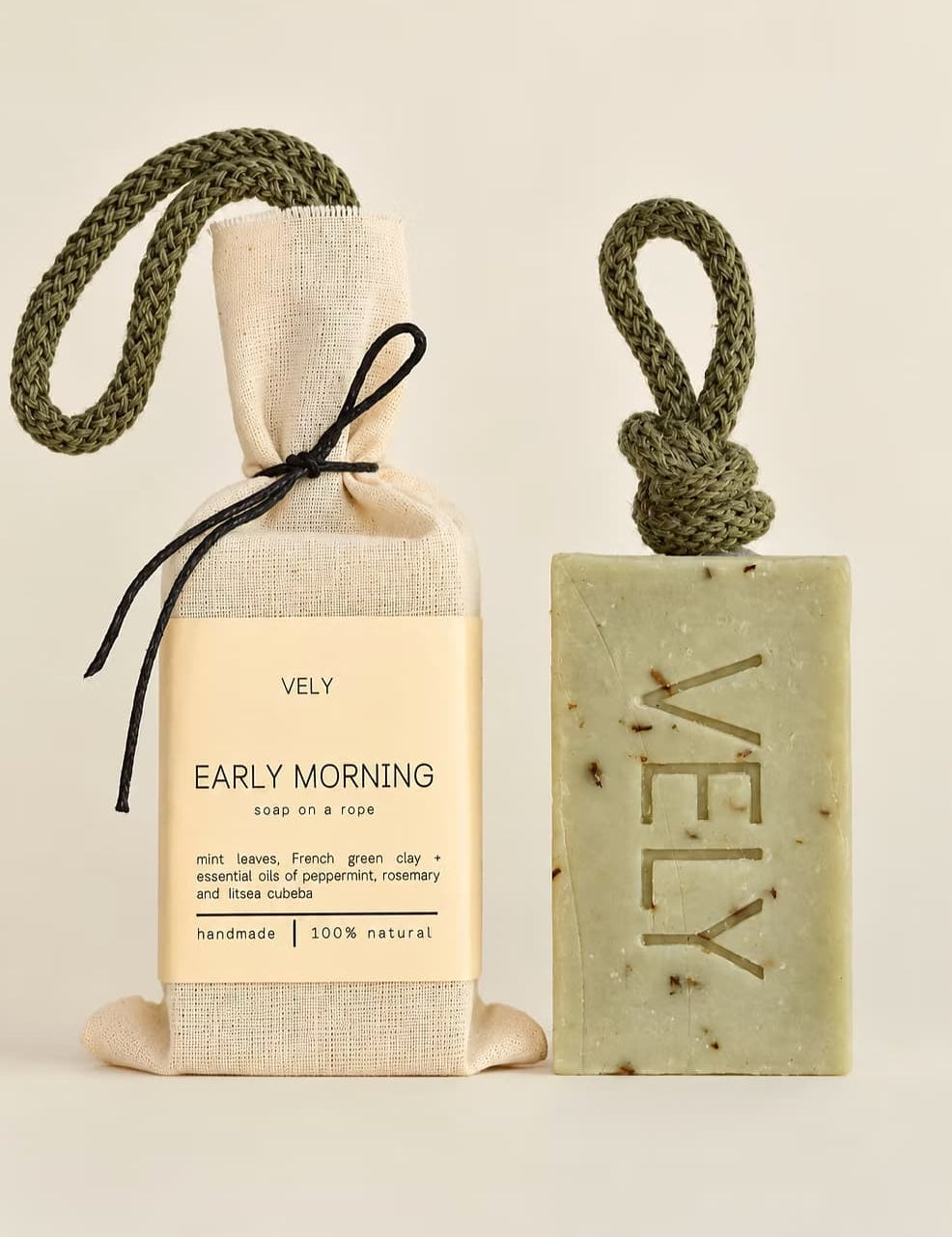 A VELY soap gift set featuring a rectangular speckled green soap bar with a rope loop and a beige fabric-wrapped soap bag tied with a black ribbon, both labeled 'VELY EARLY MORNING'.