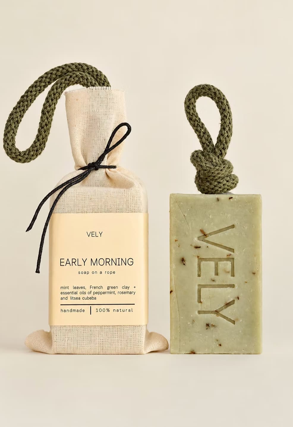 A VELY soap gift set featuring a rectangular speckled green soap bar with a rope loop and a beige fabric-wrapped soap bag tied with a black ribbon, both labeled 'VELY EARLY MORNING'.