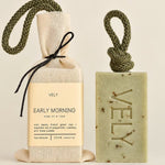A VELY soap gift set featuring a rectangular speckled green soap bar with a rope loop and a beige fabric-wrapped soap bag tied with a black ribbon, both labeled 'VELY EARLY MORNING'.
