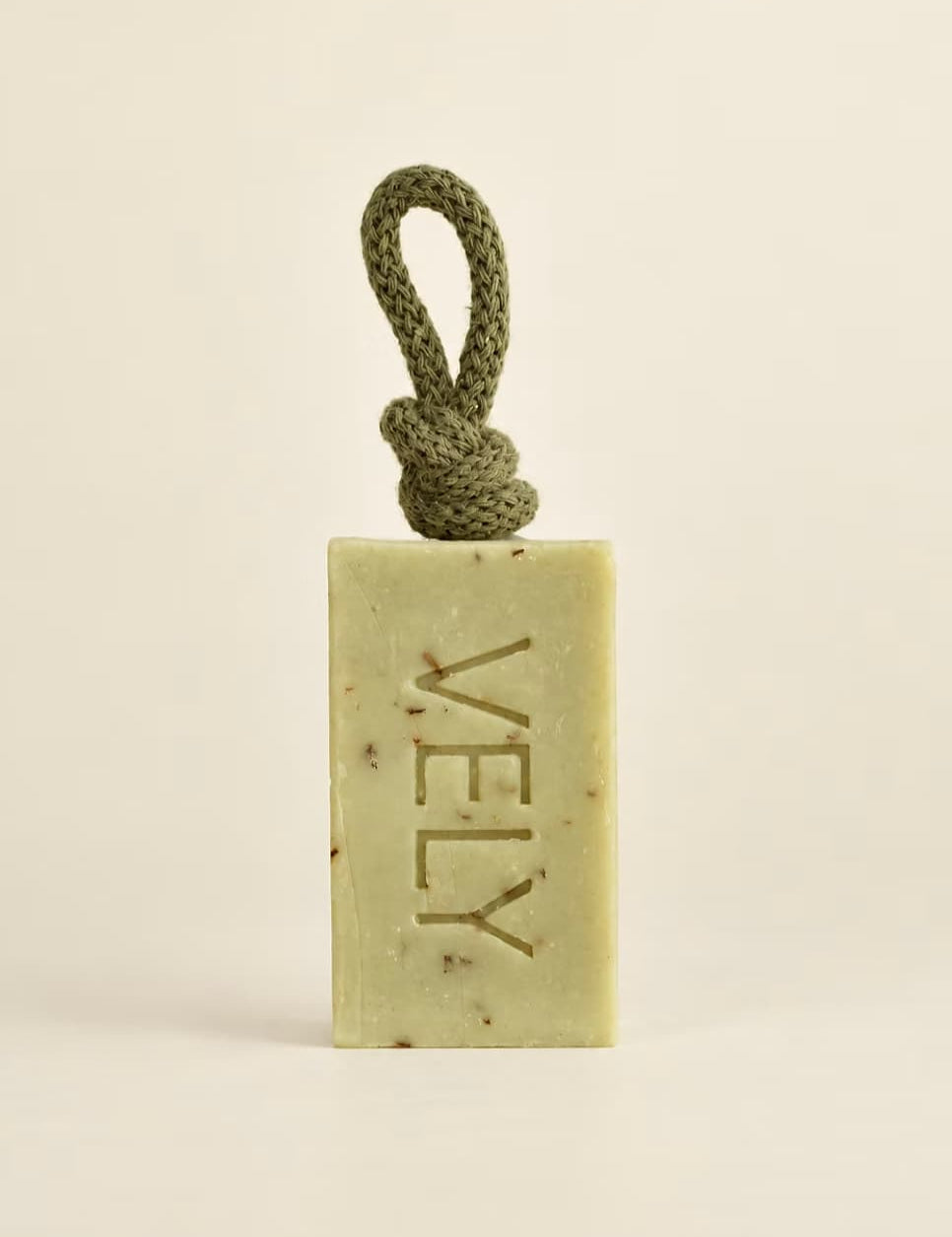 A rectangular bar of VELY soap in a pale yellow-green hue with embedded botanical specks, featuring a knotted olive green cord loop at the top. The brand name 'VELY' is embossed vertically on the front.