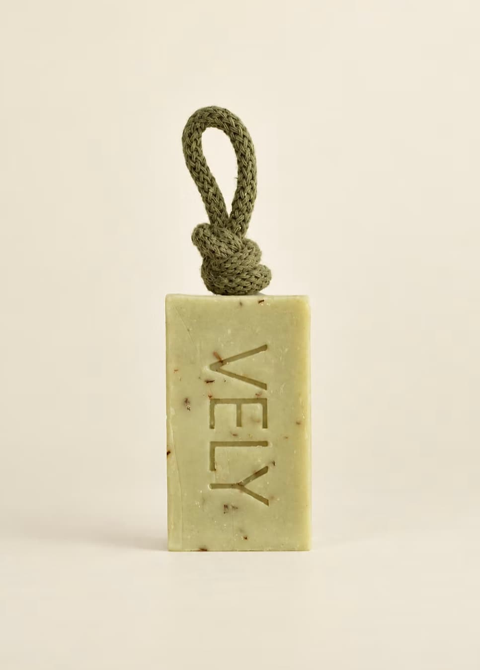 A rectangular bar of VELY soap in a pale yellow-green hue with embedded botanical specks, featuring a knotted olive green cord loop at the top. The brand name 'VELY' is embossed vertically on the front.
