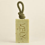 A rectangular bar of VELY soap in a pale yellow-green hue with embedded botanical specks, featuring a knotted olive green cord loop at the top. The brand name 'VELY' is embossed vertically on the front.