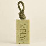 A rectangular bar of VELY soap in a pale yellow-green hue with embedded botanical specks, featuring a knotted olive green cord loop at the top. The brand name 'VELY' is embossed vertically on the front.
