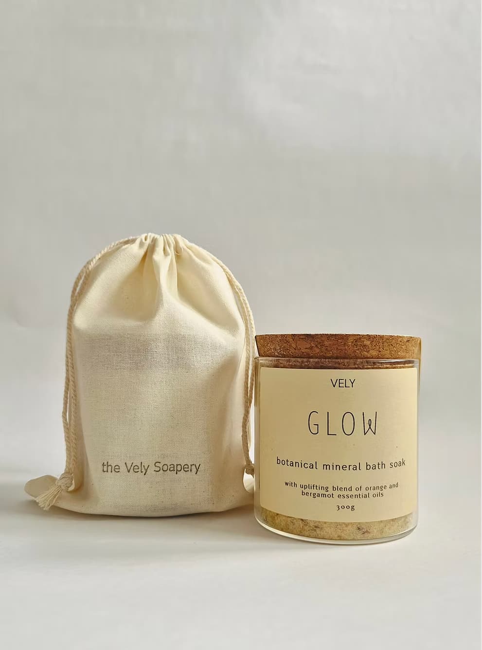 A glass jar of VELY Glow botanical mineral bath soak with a cork lid and cream label, shown next to a matching cream drawstring pouch printed with 'the Vely Scapery' on a light gray background.