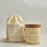 A glass jar of VELY Glow botanical mineral bath soak with a cork lid and cream label, shown next to a matching cream drawstring pouch printed with 'the Vely Scapery' on a light gray background.