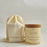 A glass jar of VELY Glow botanical mineral bath soak with a cork lid and cream label, shown next to a matching cream drawstring pouch printed with 'the Vely Scapery' on a light gray background.