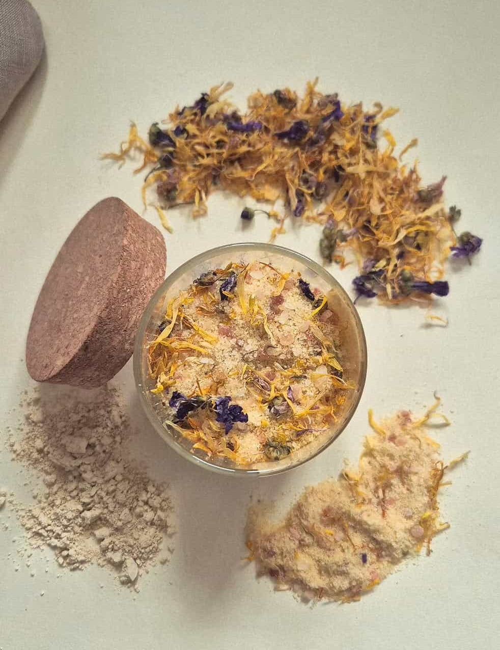 A small clear jar filled with a beige bath or body powder topped with dried yellow and purple flowers, shown on a light neutral background with loose powder, a round wooden scoop, and scattered dried botanicals around it.