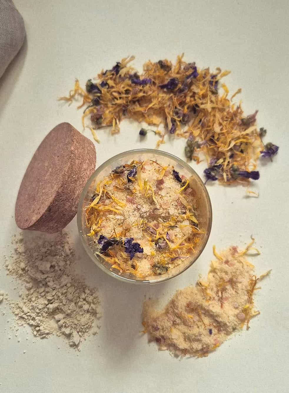 A small clear jar filled with a beige bath or body powder topped with dried yellow and purple flowers, shown on a light neutral background with loose powder, a round wooden scoop, and scattered dried botanicals around it.