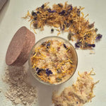 A small clear jar filled with a beige bath or body powder topped with dried yellow and purple flowers, shown on a light neutral background with loose powder, a round wooden scoop, and scattered dried botanicals around it.