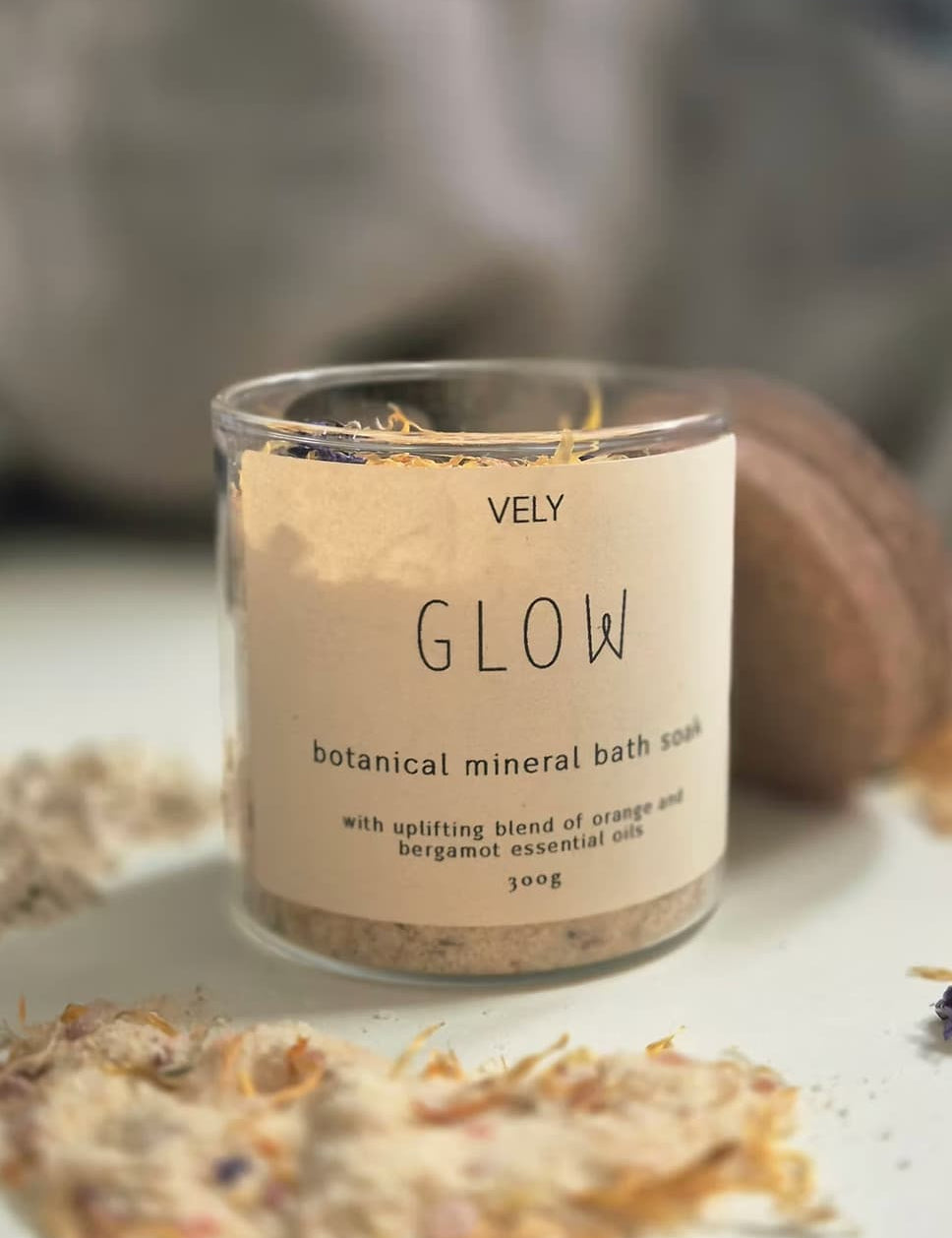 A clear glass jar of VELY Glow botanical mineral bath soak with a beige label, set on a white surface with dried botanicals scattered around it.