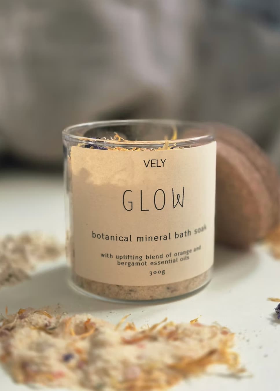 A clear glass jar of VELY Glow botanical mineral bath soak with a beige label, set on a white surface with dried botanicals scattered around it.