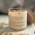A clear glass jar of VELY Glow botanical mineral bath soak with a beige label, set on a white surface with dried botanicals scattered around it.