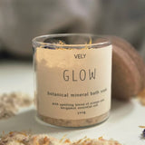 A clear glass jar of VELY Glow botanical mineral bath soak with a beige label, set on a white surface with dried botanicals scattered around it.