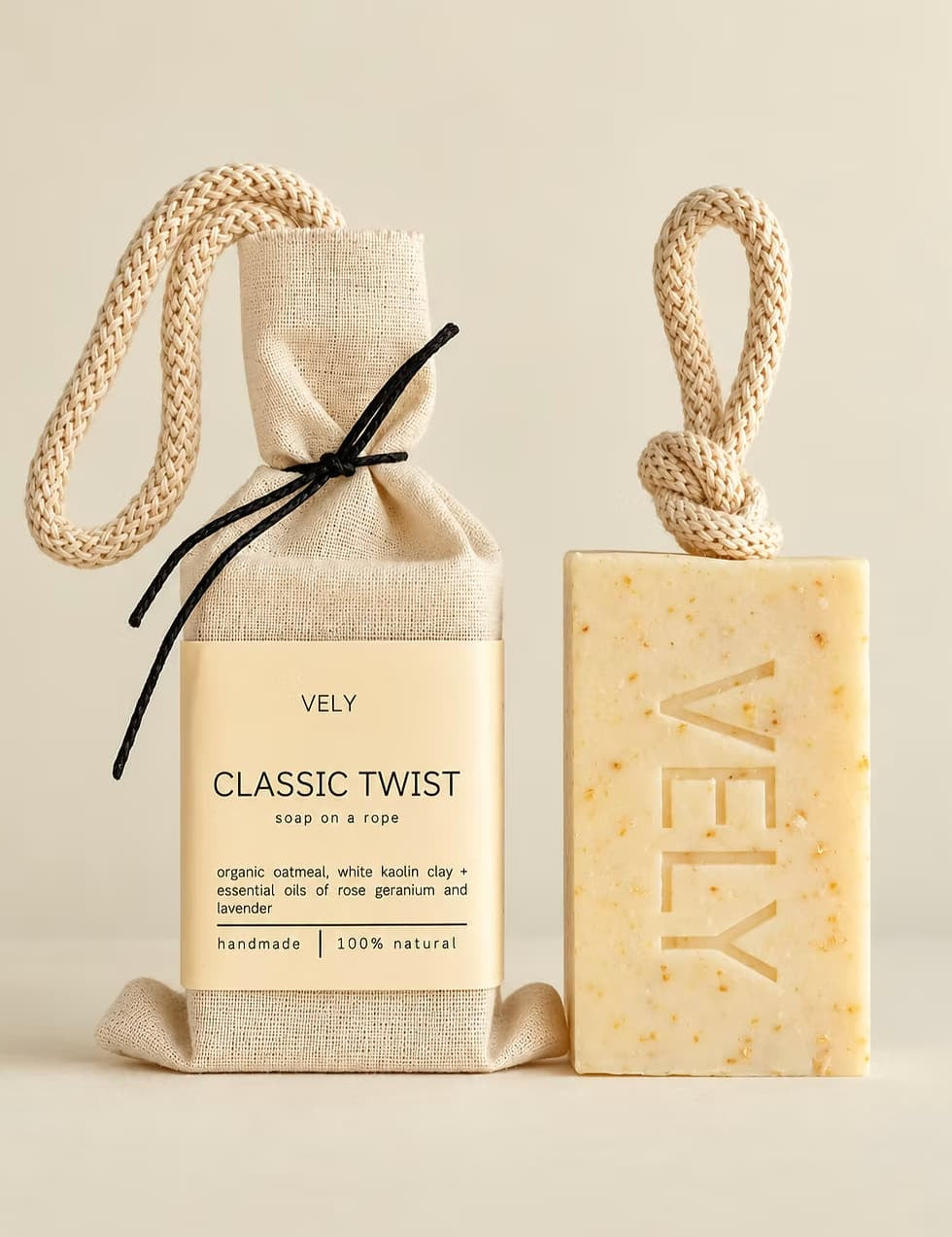 Vely Classic Twist soap shown as a packaged cloth soap bag next to a bar of soap on a warm beige background. The soap bar is pale yellow with tiny speckles and embossed with the brand name 'VELY'.