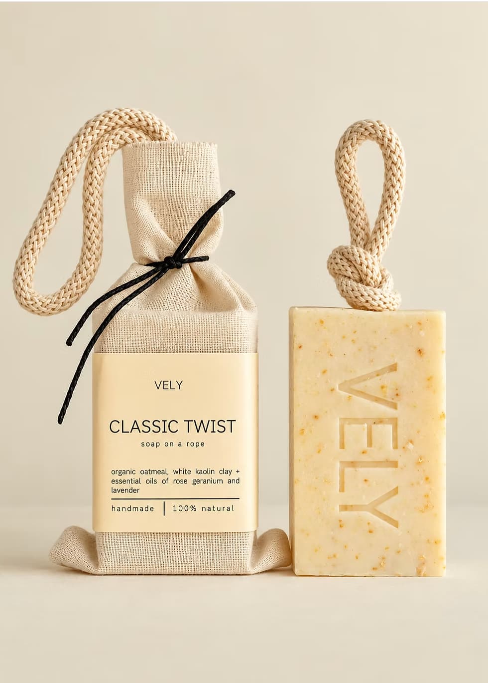 Vely Classic Twist soap shown as a packaged cloth soap bag next to a bar of soap on a warm beige background. The soap bar is pale yellow with tiny speckles and embossed with the brand name 'VELY'.