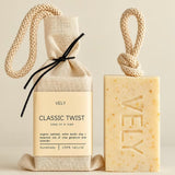 Vely Classic Twist soap shown as a packaged cloth soap bag next to a bar of soap on a warm beige background. The soap bar is pale yellow with tiny speckles and embossed with the brand name 'VELY'.