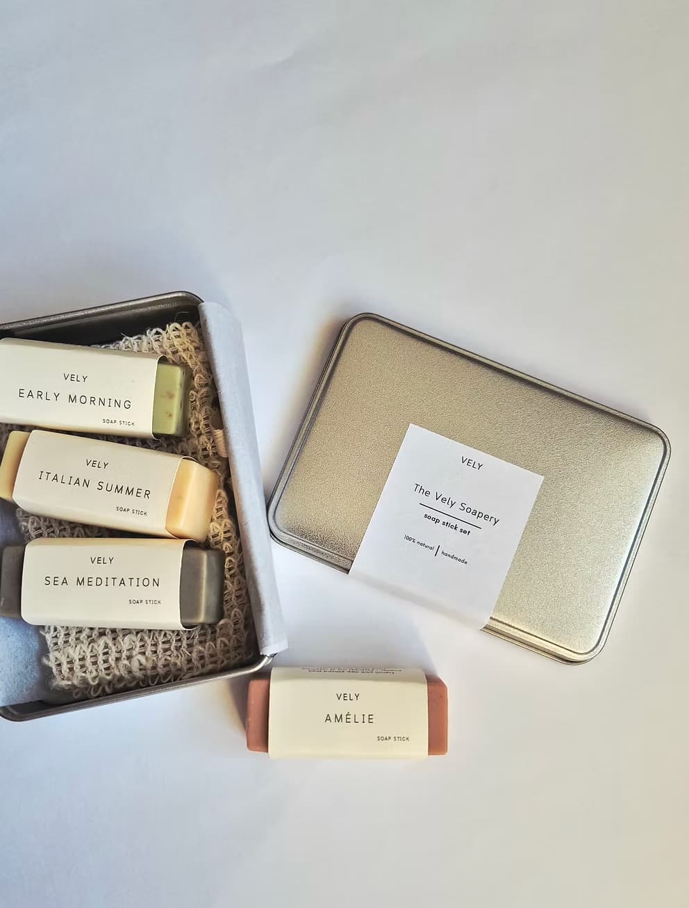 Vely soap gift set arranged on a white background, including an open tin with four wrapped bars, a closed metallic tin with a paper label, and one soap bar placed in front. Visible labels show the brand Vely and soap names Early Morning, Italian Summer, Sea Meditation, and Amelie.