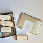 Vely soap gift set arranged on a white background, including an open tin with four wrapped bars, a closed metallic tin with a paper label, and one soap bar placed in front. Visible labels show the brand Vely and soap names Early Morning, Italian Summer, Sea Meditation, and Amelie.