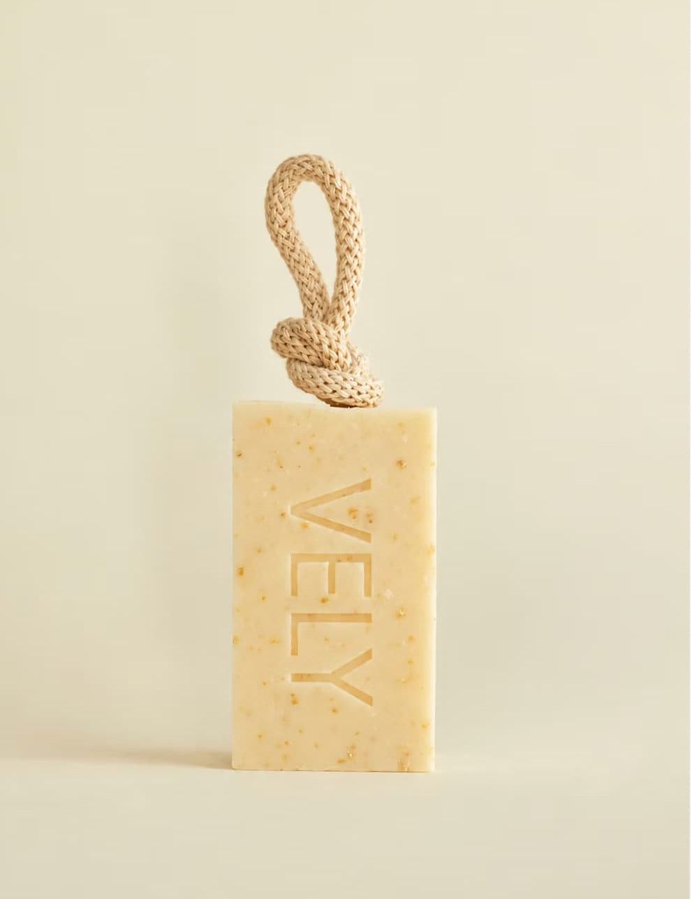 A rectangular bar of VELY soap in a warm cream-beige color with tiny speckles, featuring a knotted natural-fiber loop at the top. The brand name 'VELY' is embossed vertically on the front.