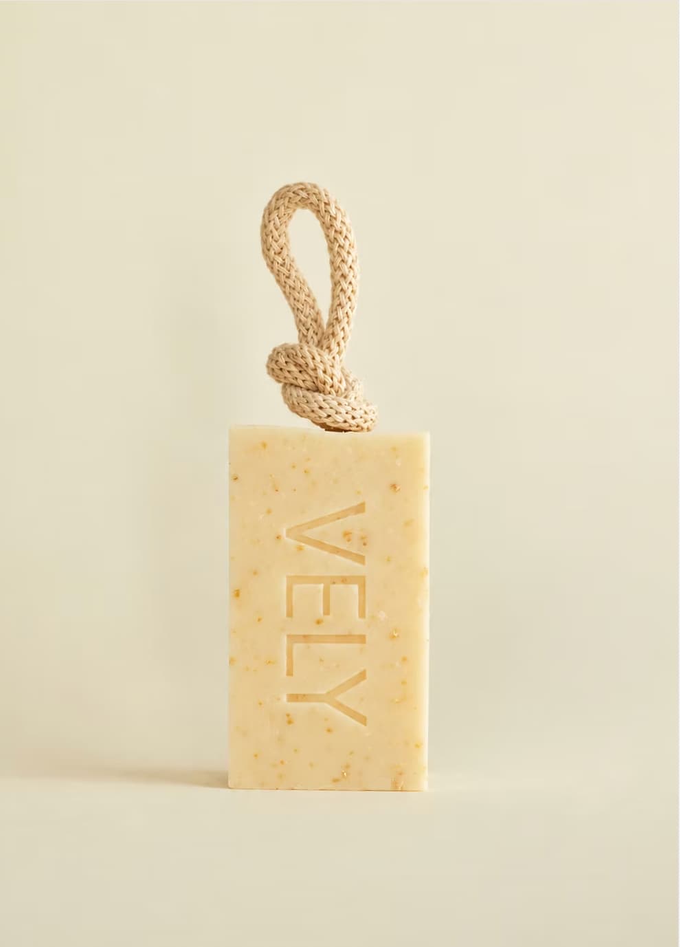 A rectangular bar of VELY soap in a warm cream-beige color with tiny speckles, featuring a knotted natural-fiber loop at the top. The brand name 'VELY' is embossed vertically on the front.