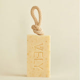 A rectangular bar of VELY soap in a warm cream-beige color with tiny speckles, featuring a knotted natural-fiber loop at the top. The brand name 'VELY' is embossed vertically on the front.