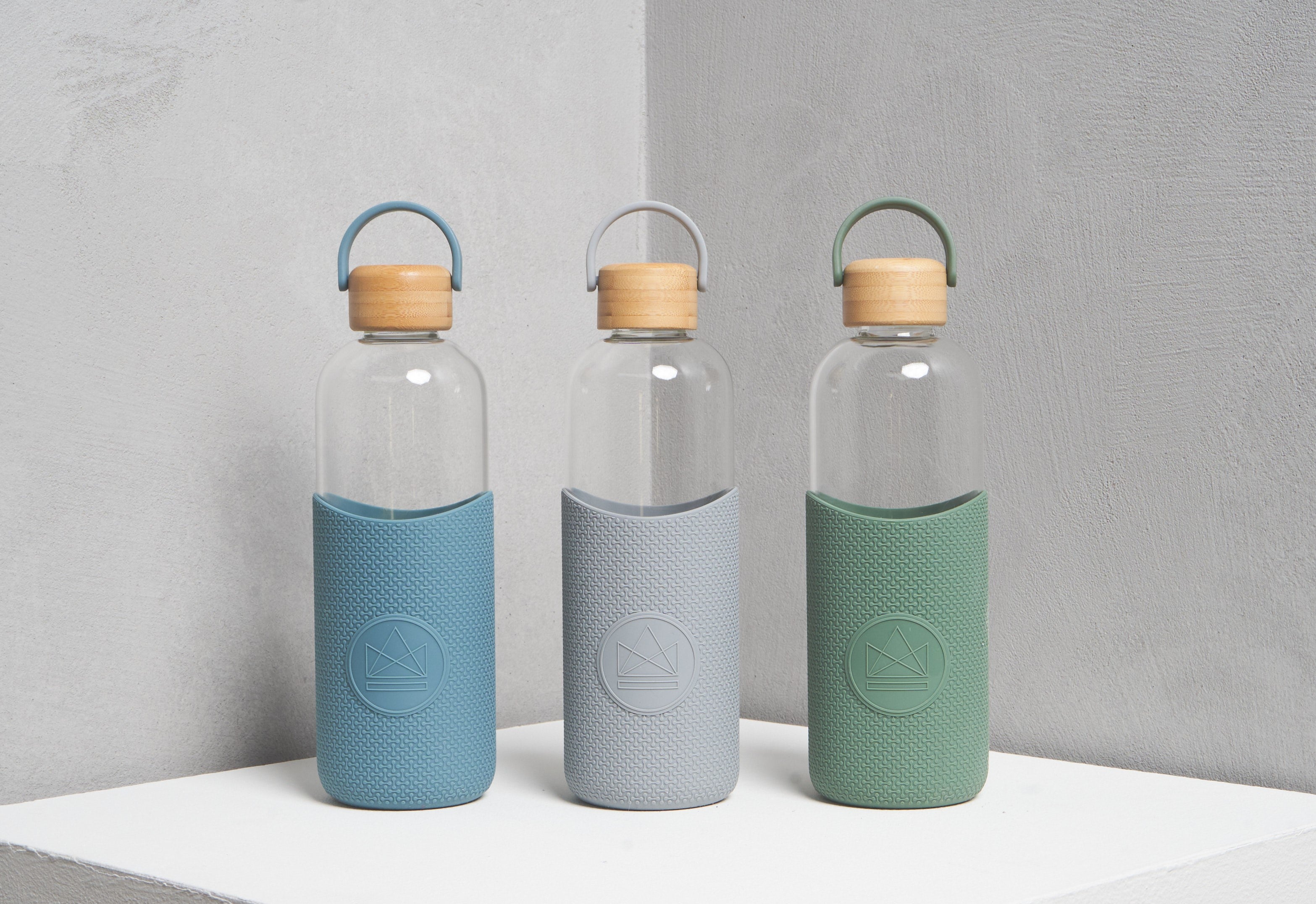 Lifestyle photo of three 1L reusable glass water bottles with silicone sleeves and bamboo lids in assorted colours, sustainable takeaway merch and eco-friendly corporate gifts