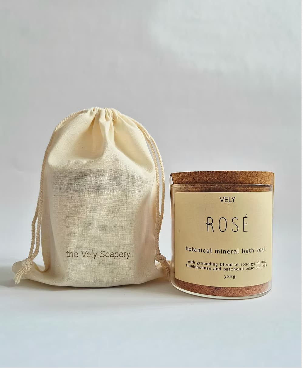 A jar of Vely Rosé botanical mineral bath soak next to a cream-colored drawstring pouch with 'the Vely Soapery' printed on it, set against a plain light gray background.