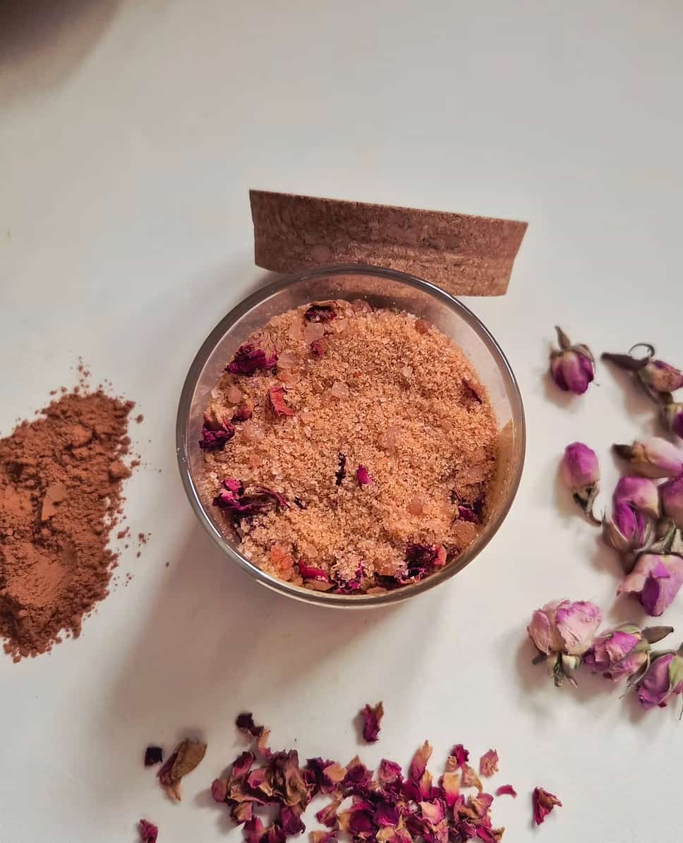 A clear glass bowl filled with a light brown exfoliating scrub, surrounded by loose rose petals, a small pile of brown powder, and a piece of cork on a white surface.