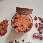 A clear glass bowl filled with a light brown exfoliating scrub, surrounded by loose rose petals, a small pile of brown powder, and a piece of cork on a white surface.