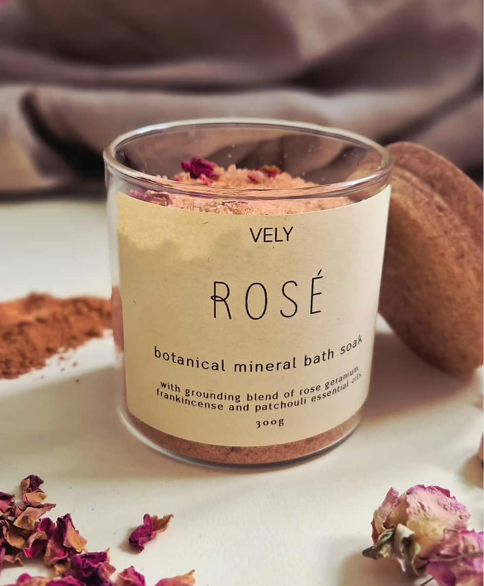 A glass jar of VELY ROSÉ botanical mineral bath soak with a beige label, styled on a white surface with dried rose petals, loose powder, and a soft fabric background.