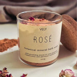 A glass jar of VELY ROSÉ botanical mineral bath soak with a beige label, styled on a white surface with dried rose petals, loose powder, and a soft fabric background.