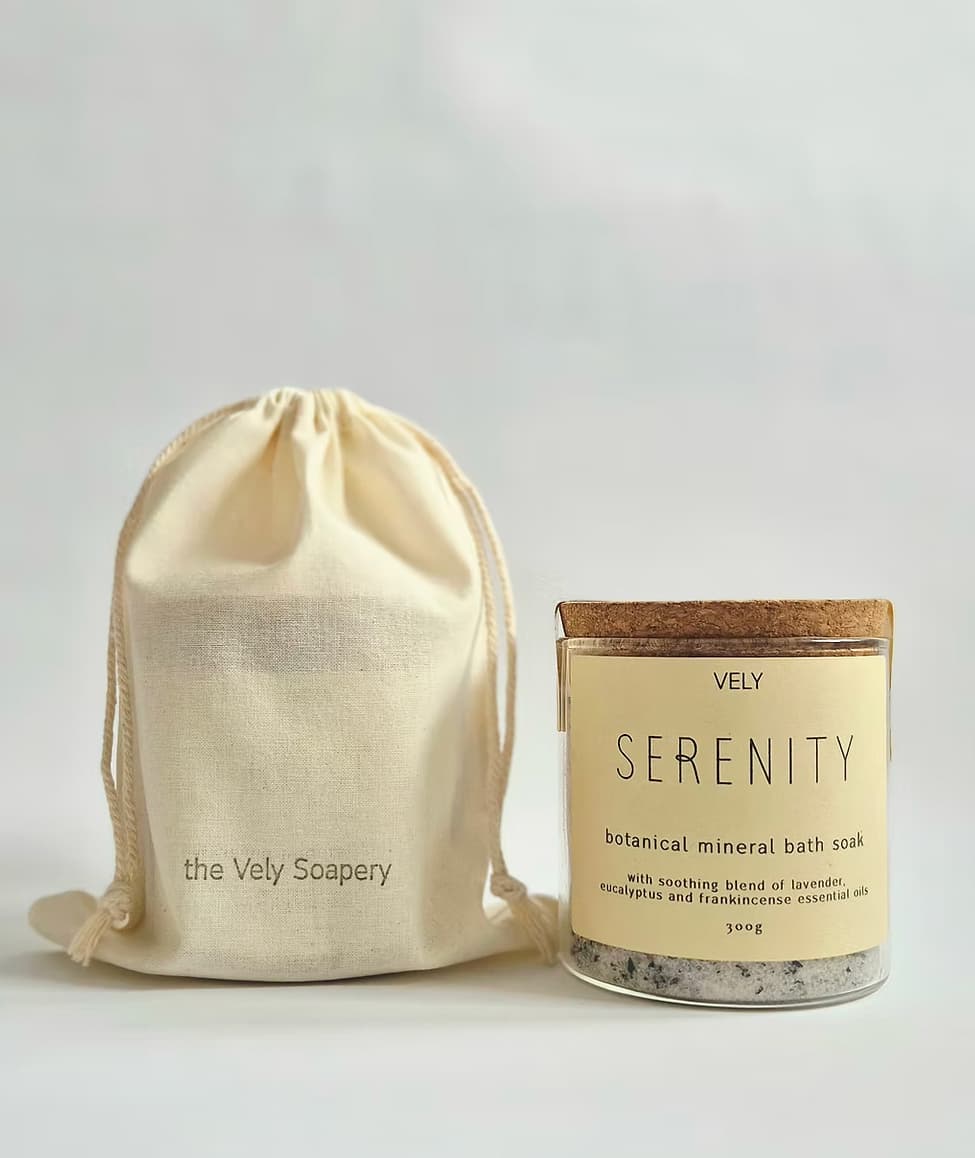 A glass jar of Vely Serenity botanical mineral bath soak with a cork lid and cream label, shown next to a matching cream drawstring pouch printed with 'the Vely Scapery' on a light gray background.