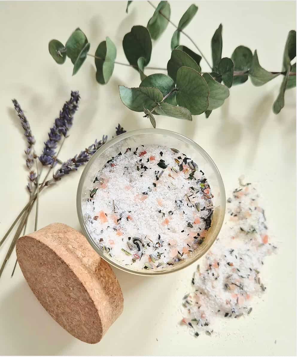 A clear glass jar filled with white bath salts mixed with pink and dark botanical pieces, shown with a cork lid beside it on a light cream background, with eucalyptus leaves and lavender sprigs as styling props.