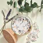 A clear glass jar filled with white bath salts mixed with pink and dark botanical pieces, shown with a cork lid beside it on a light cream background, with eucalyptus leaves and lavender sprigs as styling props.