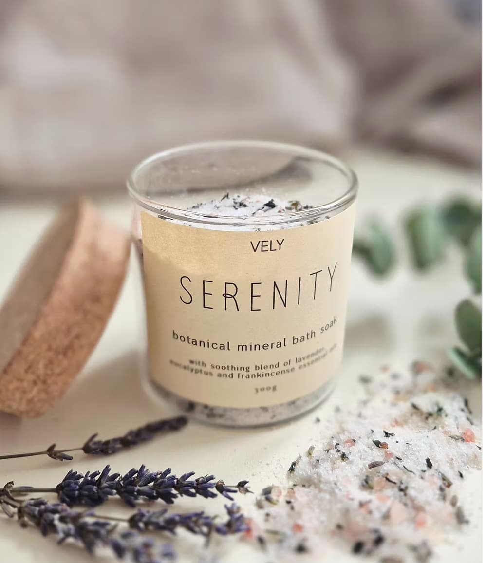 A glass jar of VELY SERENITY botanical mineral bath soak with a beige label, styled on a light surface with lavender sprigs, coarse bath salts, and a cork scrubber.