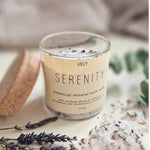 A glass jar of VELY SERENITY botanical mineral bath soak with a beige label, styled on a light surface with lavender sprigs, coarse bath salts, and a cork scrubber.