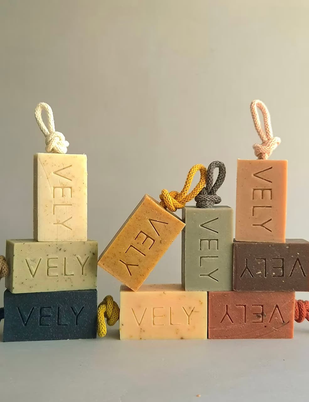 Rectangular soap bars with hanging cords, each stacked with two more soap bars behind it, on a light neutral surface against a soft gray background. The soaps are in muted earthy tones: cream, tan, dark blue-gray, sage green, peach, and terracotta, with the brand name VELY embossed on each bar.