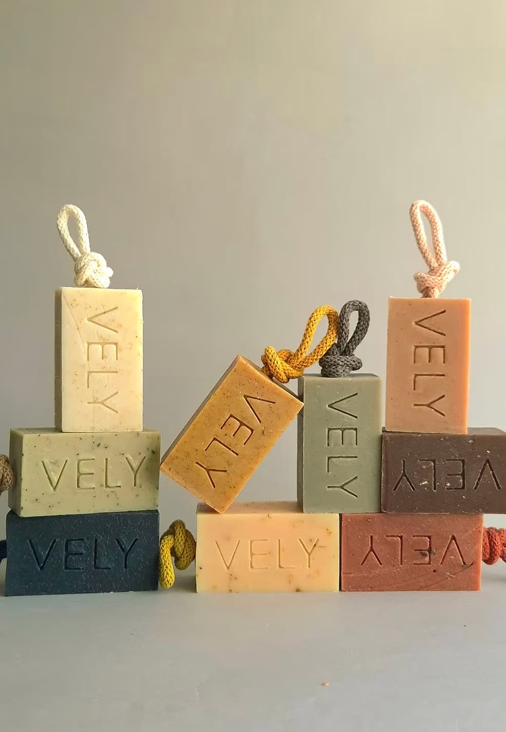 Rectangular soap bars with hanging cords, each stacked with two more soap bars behind it, on a light neutral surface against a soft gray background. The soaps are in muted earthy tones: cream, tan, dark blue-gray, sage green, peach, and terracotta, with the brand name VELY embossed on each bar.