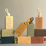 Rectangular soap bars with hanging cords, each stacked with two more soap bars behind it, on a light neutral surface against a soft gray background. The soaps are in muted earthy tones: cream, tan, dark blue-gray, sage green, peach, and terracotta, with the brand name VELY embossed on each bar.