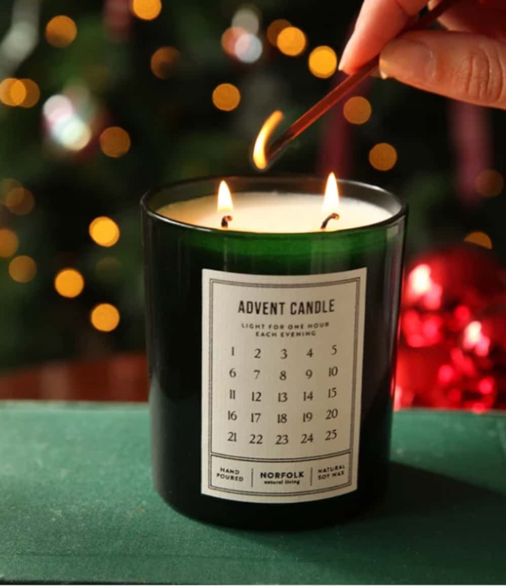 A dark green glass candle labeled 'ADVENT CANDLE' with two lit wicks, being lit with a wooden match in a festive setting with blurred Christmas lights and ornaments in the background.