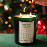A dark green glass candle labeled 'ADVENT CANDLE' with two lit wicks, being lit with a wooden match in a festive setting with blurred Christmas lights and ornaments in the background.