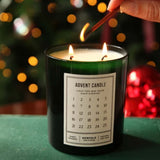 A dark green glass candle labeled 'ADVENT CANDLE' with two lit wicks, being lit with a wooden match in a festive setting with blurred Christmas lights and ornaments in the background.