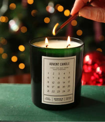 A dark green glass candle labeled 'ADVENT CANDLE' with two lit wicks, being lit with a wooden match in a festive setting with blurred Christmas lights and ornaments in the background.