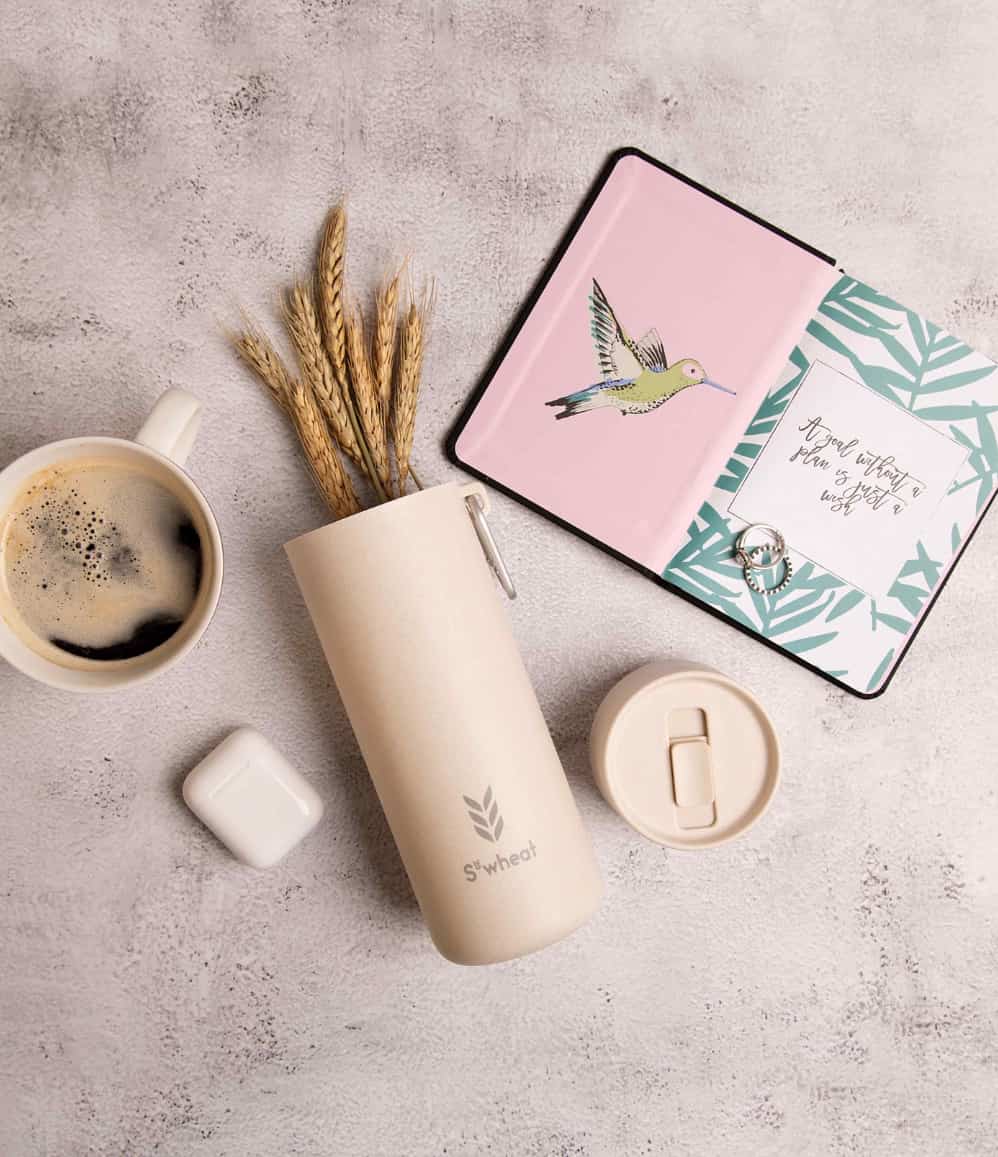 Plant Based Water Bottle in oat, with the S'wheat logo, styled with a coffee cup, dried wheat and a notebook
