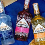 Three bottles of Warner's brand gin arranged on a blue fabric background: a clear bottle labeled Congratulations, a dark bottle labeled Thank You with pink label, and a golden bottle labeled Thank You with orange label.