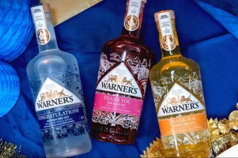 Three bottles of Warner's brand gin arranged on a blue fabric background: a clear bottle labeled Congratulations, a dark bottle labeled Thank You with pink label, and a golden bottle labeled Thank You with orange label.