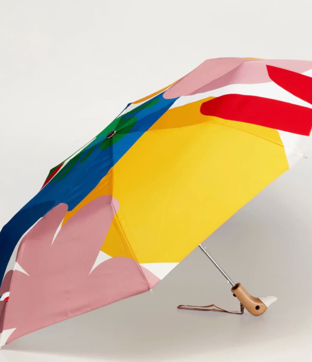 A compact folding umbrella with a bold, abstract multicolor print featuring large blocks of yellow, pink, blue, red, and white, shown open on a light gray background.