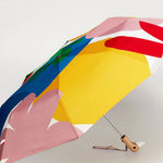 A compact folding umbrella with a bold, abstract multicolor print featuring large blocks of yellow, pink, blue, red, and white, shown open on a light gray background.