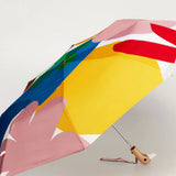 A compact folding umbrella with a bold, abstract multicolor print featuring large blocks of yellow, pink, blue, red, and white, shown open on a light gray background.